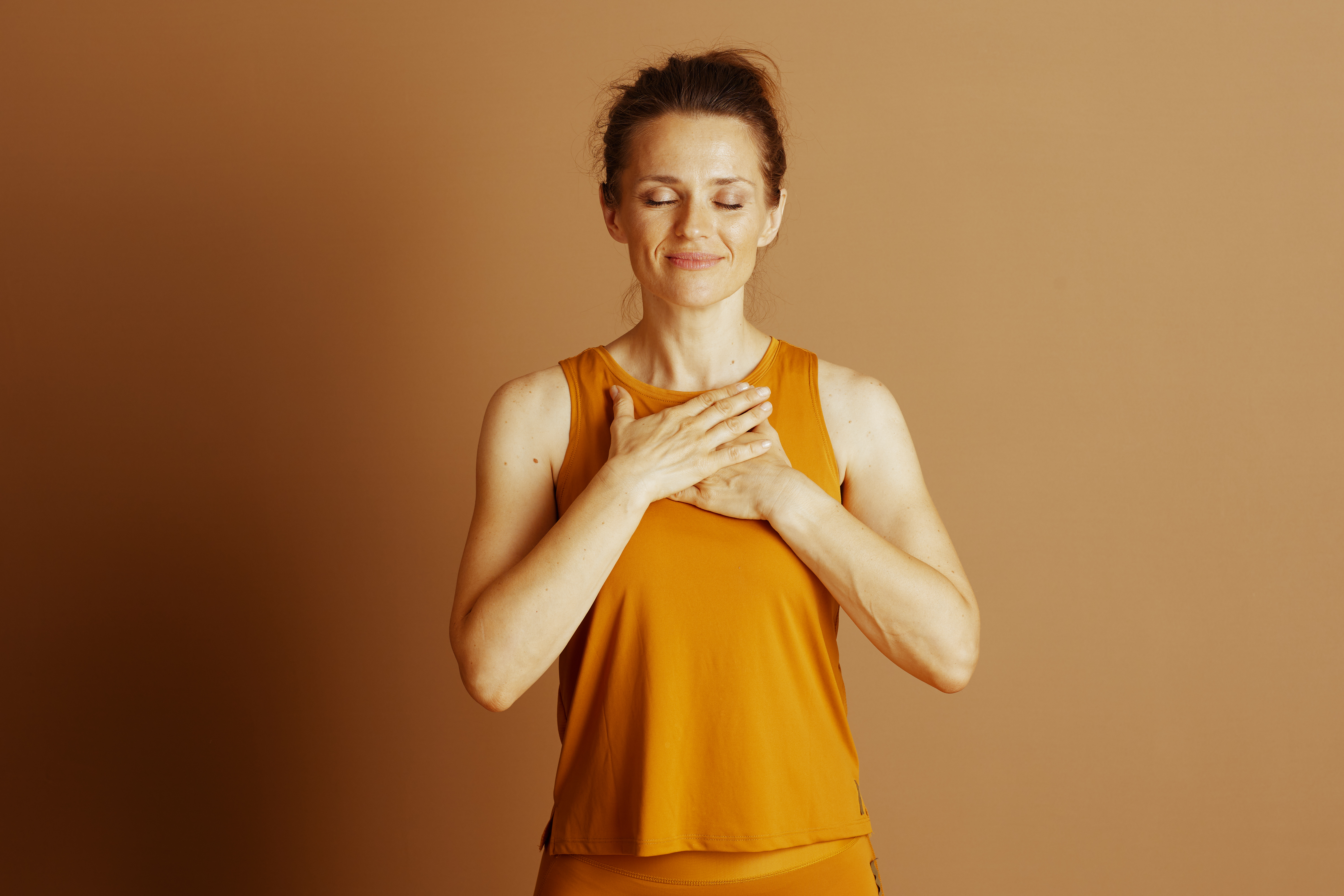 A woman with closed eyes and gentle smile places hands over chest, evoking meditative or thankful pose. Her orange sleeveless top against warm brown background suggests serenity and self-care